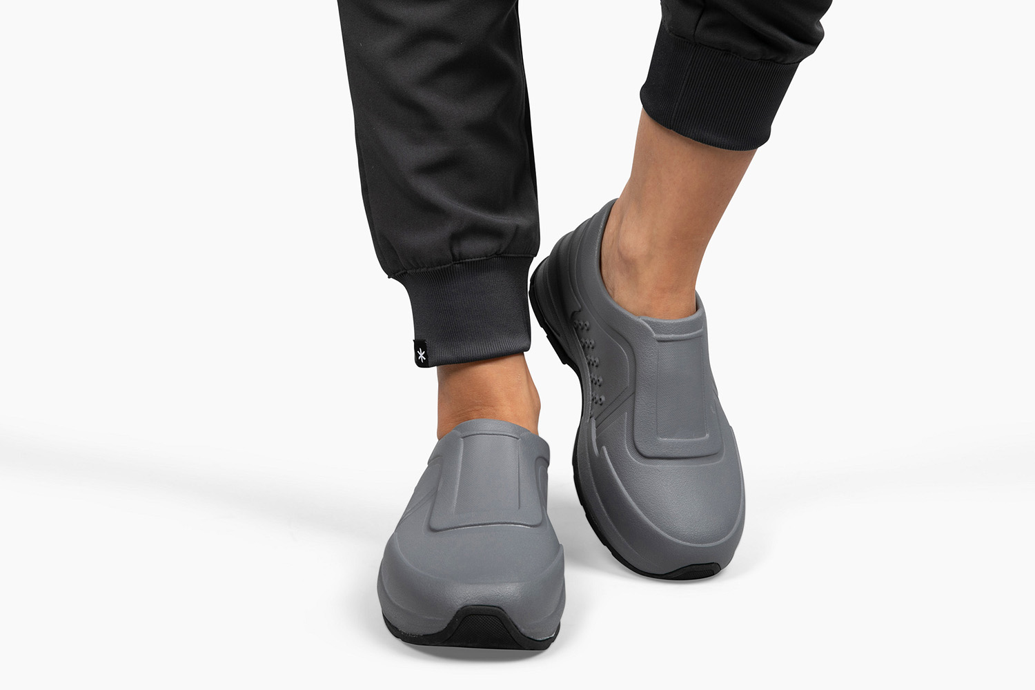 Gray slip-on shoes worn by a person dressed in black pants, walking against a white background.