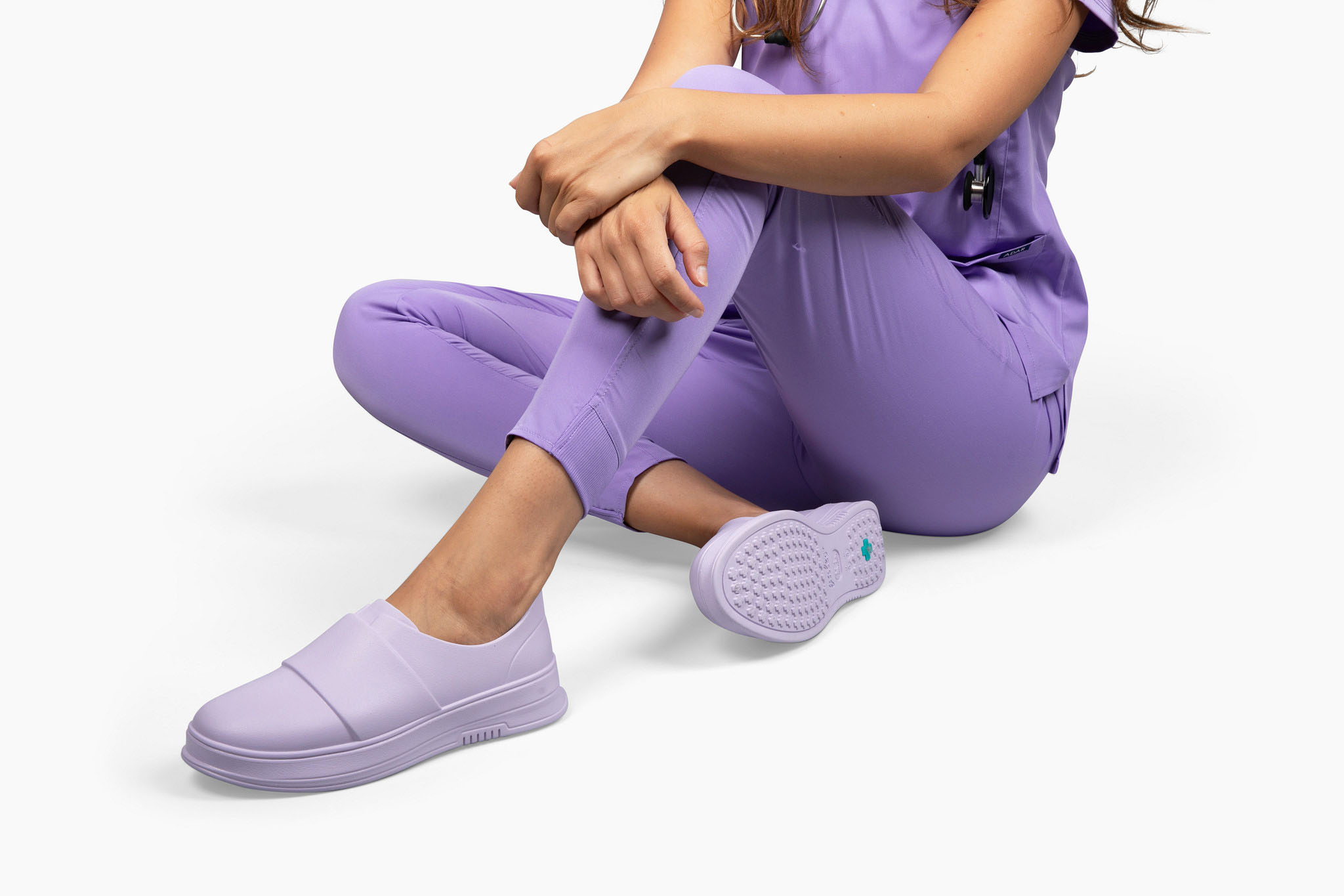 Light purple slip-on shoes worn by a person dressed in matching scrubs, sitting on a white surface.